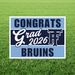 Rectangular yard sign with light blue and dark navy sections reading "CONGRATS Grad 2026 BRUINS" in white text with a bruins mascot icon, displayed on grass.