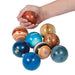 Hand holding a red metallic stress ball above a collection of 11 colorful textured balls in shades of blue, gold, bronze, and teal arranged below.
