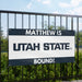 Promotional sign for Utah State University with text 'MATTHEW IS UTAH STATE BOUND!' against a natural background.