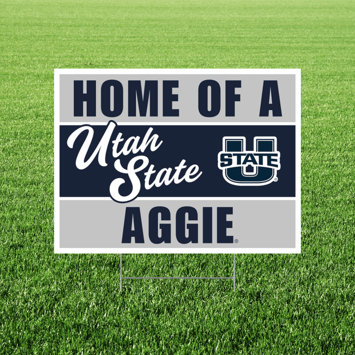 Rectangular yard sign with white border on green grass displaying "Home of a Utah State Aggie" text in navy and white with the USU State logo.