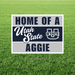 Rectangular yard sign with white border on green grass displaying "Home of a Utah State Aggie" text in navy and white with the USU State logo.