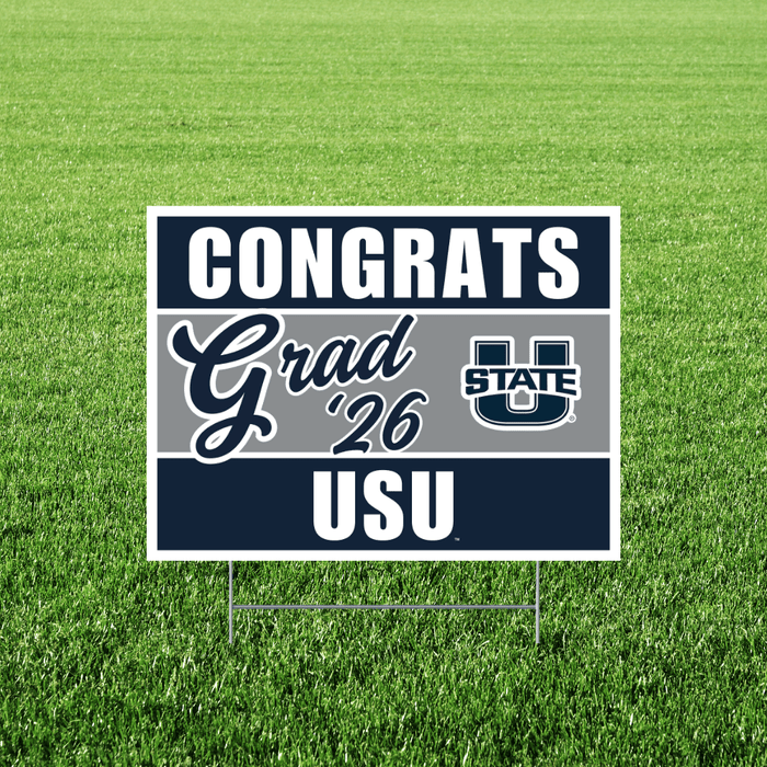 Navy and gray yard sign with white text reading "CONGRATS Grad '26 USU" and a Utah State University logo, displayed on green grass.