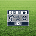 Navy and gray yard sign with white text reading "CONGRATS Grad '26 USU" and a Utah State University logo, displayed on green grass.