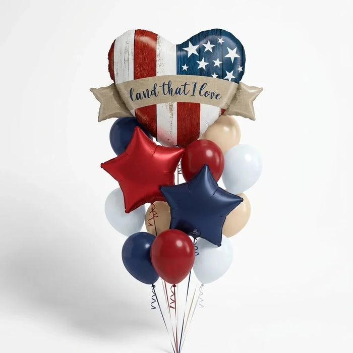 Patriotic balloon bouquet featuring a large heart-shaped centerpiece with American flag design, banner reading "land that I Love," red and navy star balloons, and coordinating red, white, and navy round balloons.