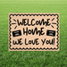 Tan corrugated yard sign with scalloped border edges displaying "WELCOME HOME WE LOVE YOU!" in black hand-painted lettering, with two small missionary name tags on either side, mounted on metal stakes in grass.