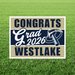Navy and tan yard sign with "CONGRATS Grad 2026 WESTLAKE" text and graduation cap illustration on green grass background.