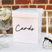 White card box with "Cards" written in black script lettering, featuring a slot at the top, displayed on a table with baby's breath flowers, a glass vase, and a white taper candle against a brick backdrop.