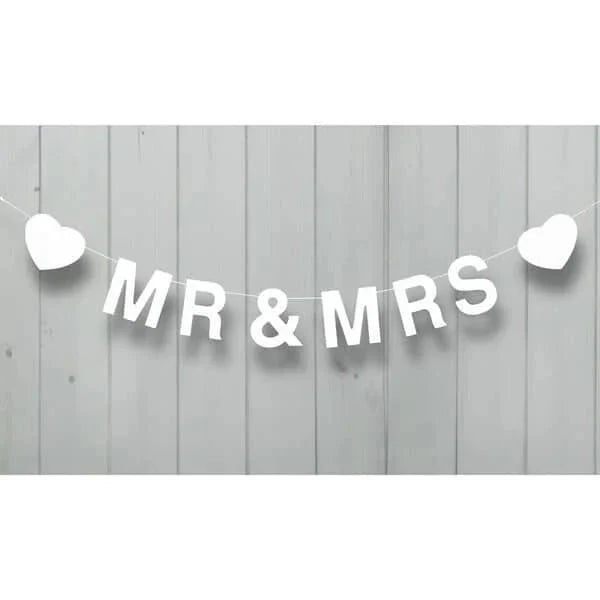 White fabric banner reading "MR & MRS" with white heart cutouts on each end, hung by string against a light gray wooden wall.