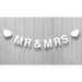 White fabric banner reading "MR & MRS" with white heart cutouts on each end, hung by string against a light gray wooden wall.
