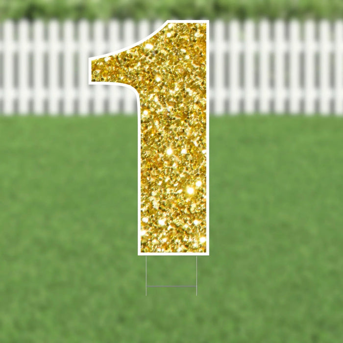 Gold glitter number 1 yard card with white border and metal stakes, displayed on green grass with white picket fence background.