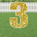 Gold glitter number 3 yard sign with white border and metal stakes, displayed on green grass with white picket fence background.