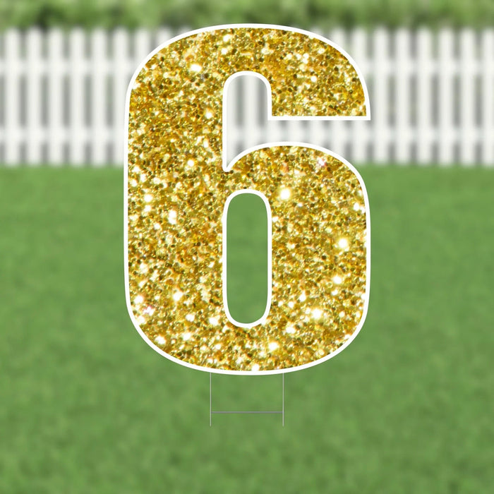 Gold glitter yard sign shaped like the number 6 with white trim, displayed on a lawn with white picket fence in the background.