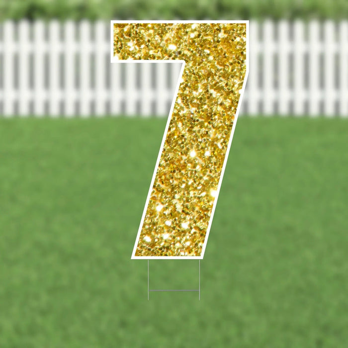Gold glitter number 7 yard sign with white border and metal stakes, displayed on a lawn with white fence in background.