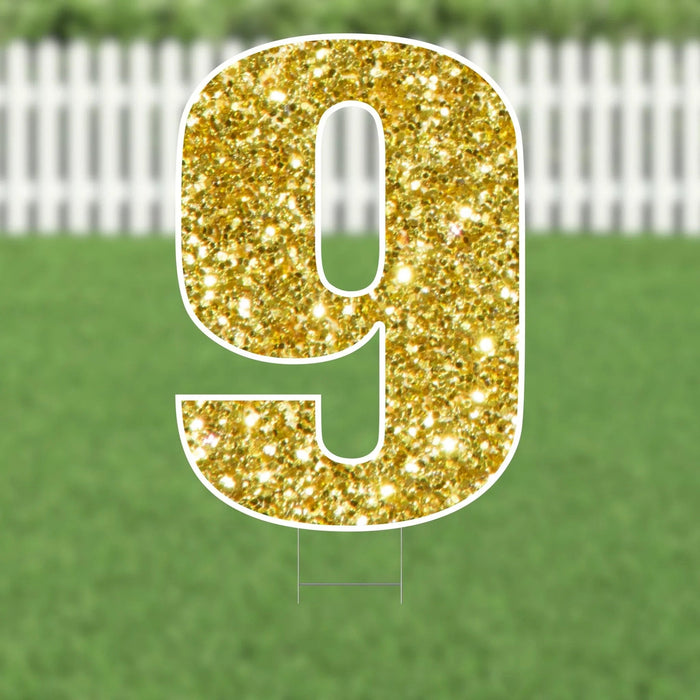Gold glitter yard sign shaped like the number 9 with a white border, displayed on green grass with a white picket fence in the background.