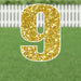 Gold glitter yard sign shaped like the number 9 with a white border, displayed on green grass with a white picket fence in the background.