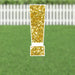 Gold glitter exclamation point yard sign with white border and metal stakes displayed on green grass with white picket fence in background.