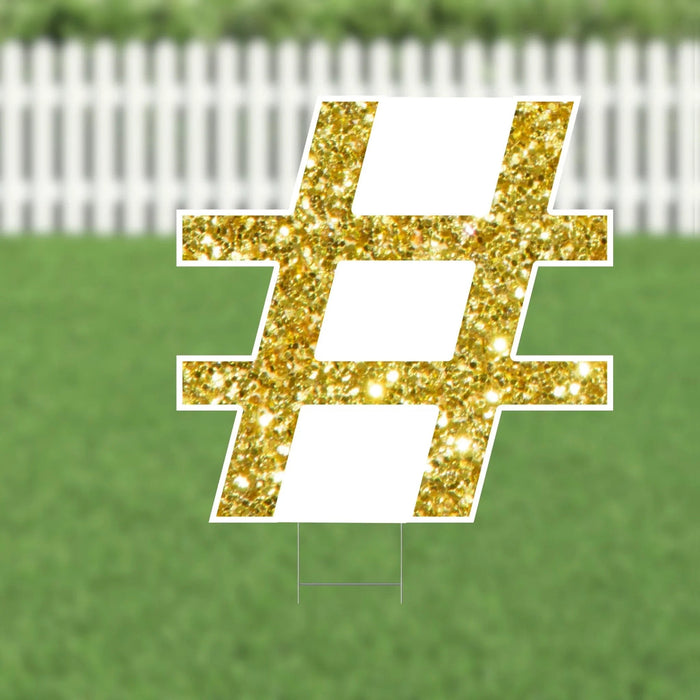Gold glitter hashtag symbol yard sign with white border and stakes, displayed on a lawn with white picket fence background.