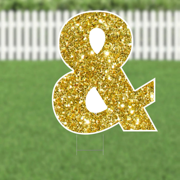 Gold glitter ampersand symbol yard sign with white border and stake legs displayed on a green lawn.