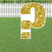 Gold glitter question mark yard sign with white border and metal stakes, displayed on green lawn with white picket fence background.