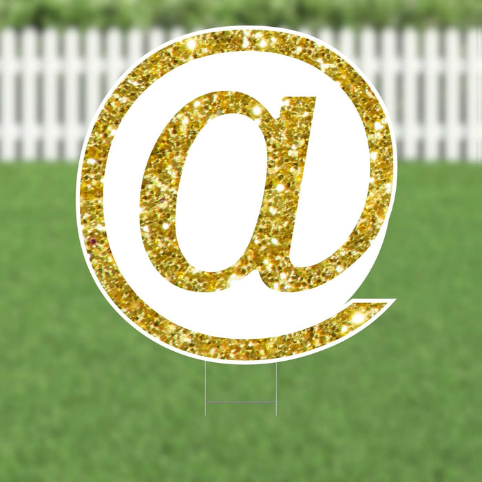 Gold glitter @ symbol yard sign with white border and metal stakes, displayed on a green lawn with white fence background.