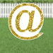 Gold glitter @ symbol yard sign with white border and metal stakes, displayed on a green lawn with white fence background.