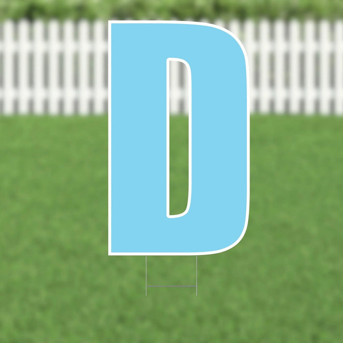 Bright blue yard sign shaped like the letter D with white border and stakes, displayed on a grass lawn with white fence in background.
