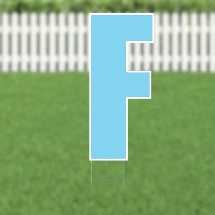 Light blue letter F yard sign with white border and metal stakes, displayed on green grass with white picket fence in background.