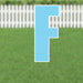 Light blue letter F yard sign with white border and metal stakes, displayed on green grass with white picket fence in background.