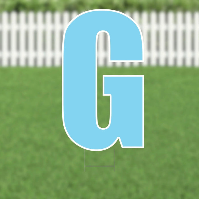 Large blue letter G yard sign with white border and metal stakes, displayed on green grass with white picket fence background.