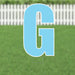 Large blue letter G yard sign with white border and metal stakes, displayed on green grass with white picket fence background.