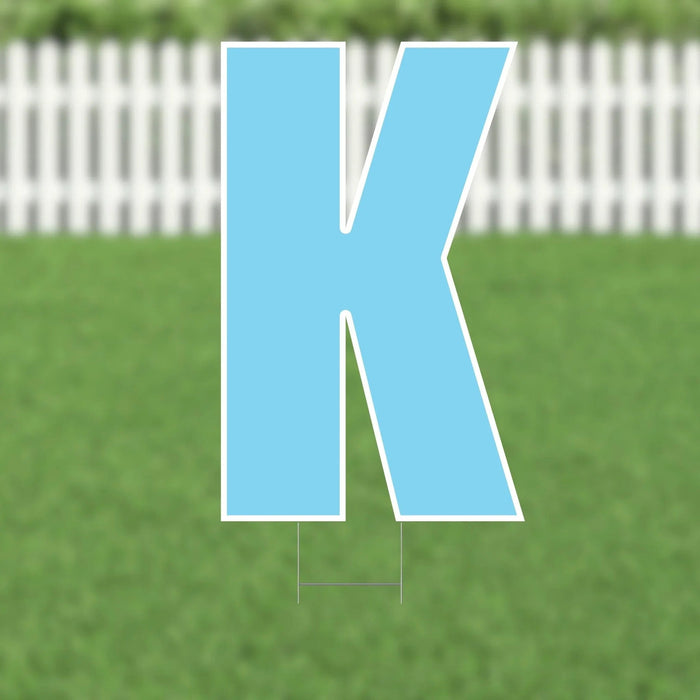 Large blue letter K yard sign with white border and metal stakes, displayed on green grass with white picket fence background.
