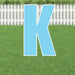 Large blue letter K yard sign with white border and metal stakes, displayed on green grass with white picket fence background.