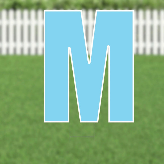 Large blue letter M yard sign with white border and metal stakes, displayed on green grass with white fence background.