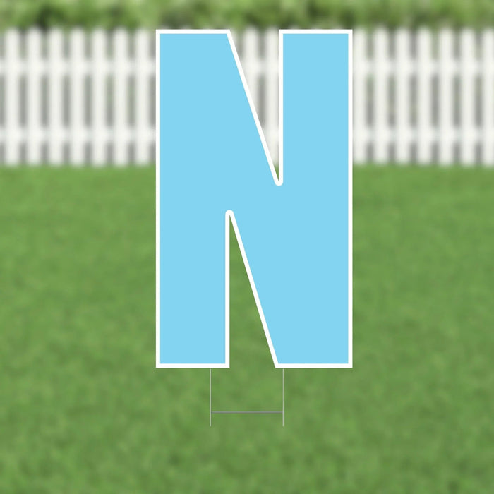 Bright blue letter N yard sign with white border and metal stakes, displayed on a lawn with white picket fence background.