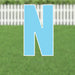 Bright blue letter N yard sign with white border and metal stakes, displayed on a lawn with white picket fence background.