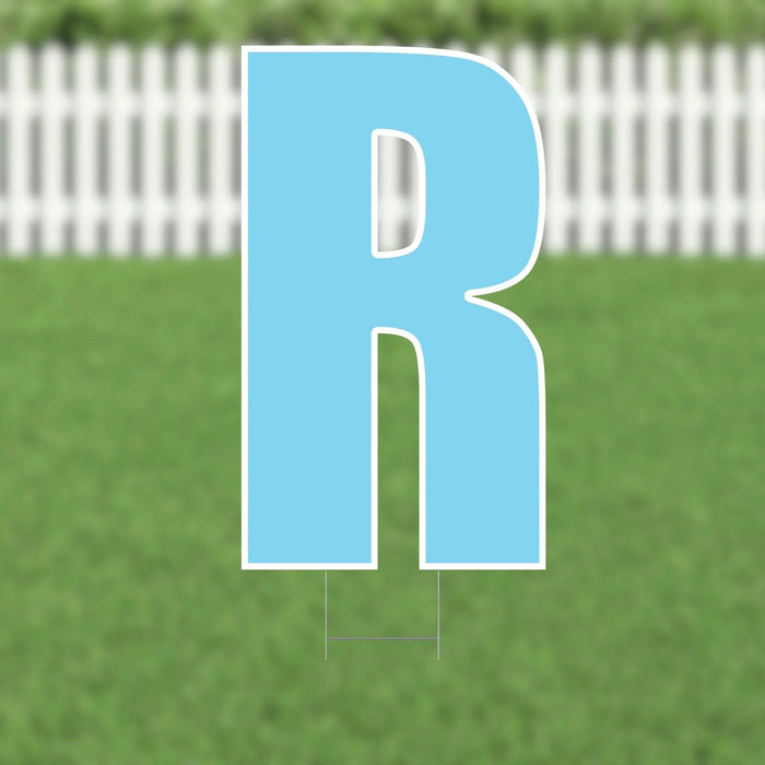 Light blue yard sign shaped like the letter R with white outline, displayed on grass with a white picket fence in the background.