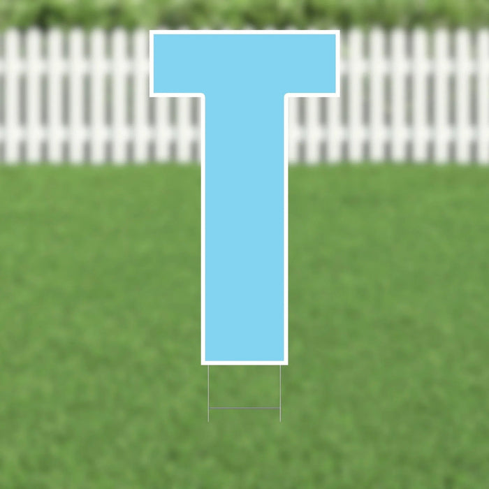 Bright blue letter T yard sign with white border and metal stakes, displayed on a grass lawn with white picket fence in background.