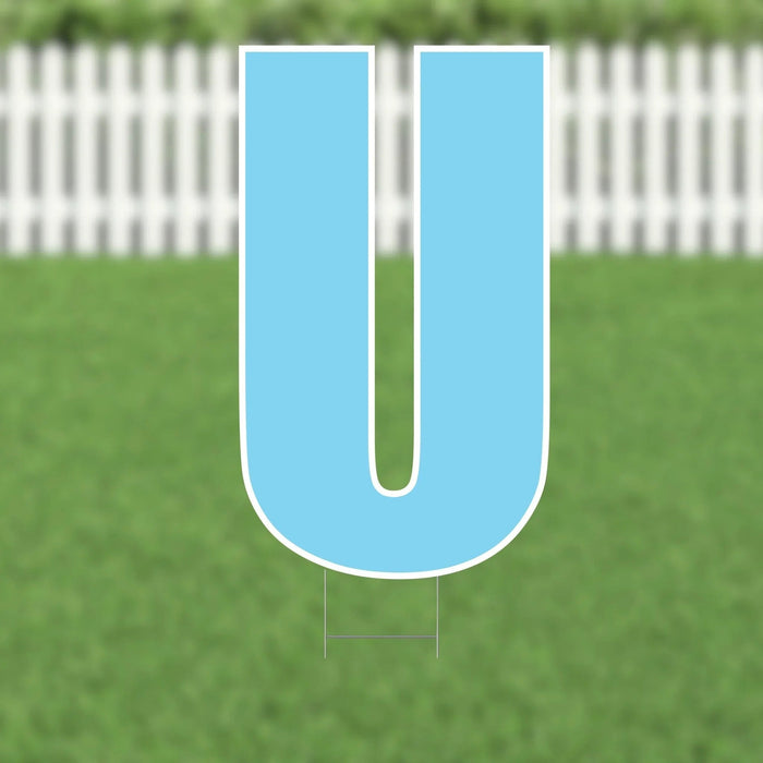 Large blue letter U yard sign with white border and metal ground stakes, displayed on green grass with white picket fence backdrop.