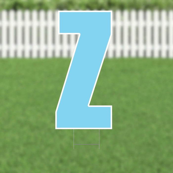Large light blue letter Z yard sign with white outline and metal stakes, displayed on a grass lawn with white picket fence background.
