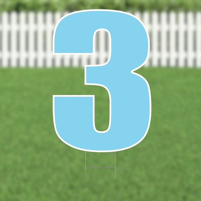 Large light blue number 3 yard sign with white outline and supporting stakes, displayed on green grass with a white picket fence background.
