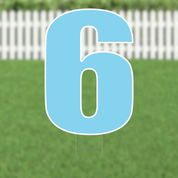 Light blue yard sign shaped as the number 6 with white outline and mounting stakes, displayed against a green lawn and white picket fence.