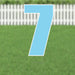 Light blue yard sign shaped like the number 7 with white border and metal stakes, displayed on green grass with white picket fence background.