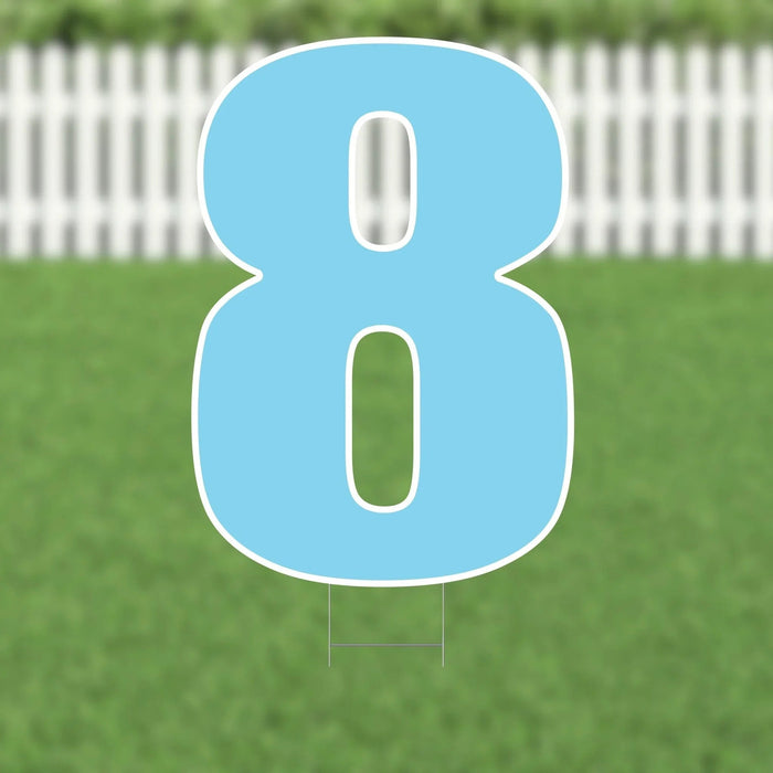 Light blue yard sign shaped like the number 8 with white outline and two support stakes, displayed on green grass with a white picket fence background.