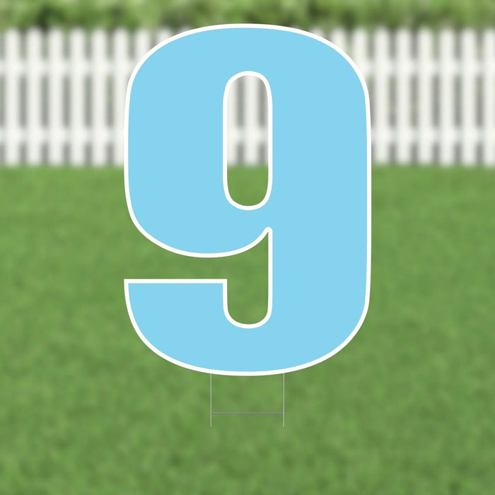 Large light blue yard sign displaying the number 9 with white outline, mounted on two metal stakes against a residential lawn and white picket fence background.
