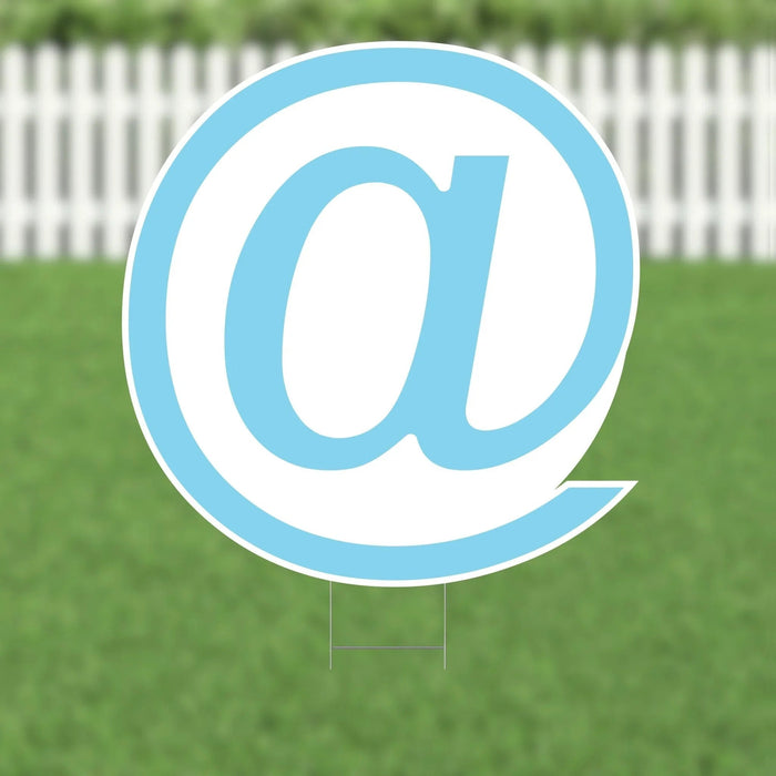 Light blue yard sign with a white @ symbol inside a circle, displayed on grass with a white picket fence in the background.
