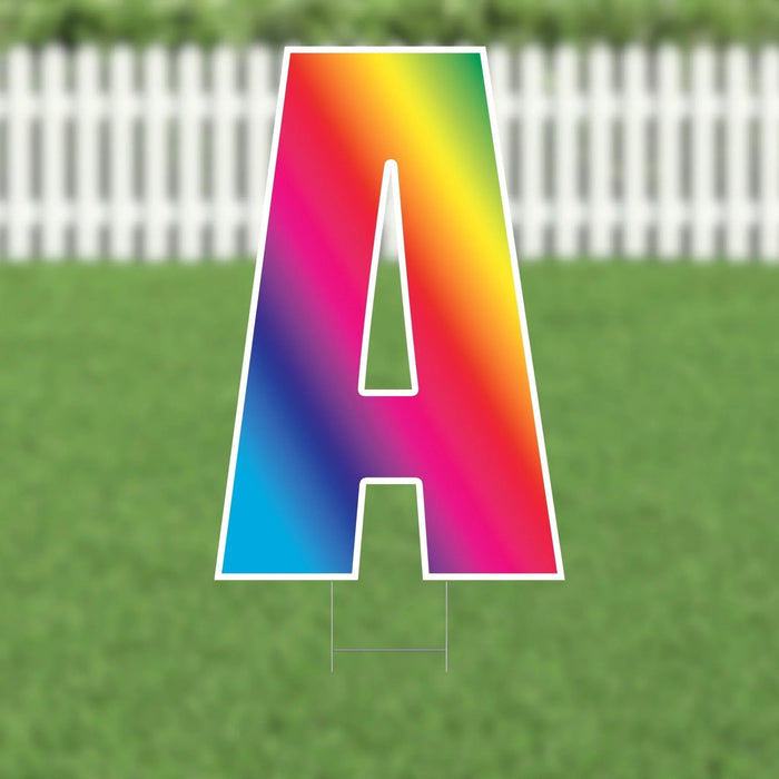 Colorful yard card sign shaped like the letter A with a rainbow gradient transitioning from blue, cyan, and purple on the left side to red, orange, and yellow on the right side, displayed in grass with a white picket fence background.