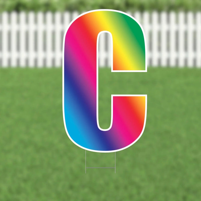 Colorful yard sign letter C with a rainbow gradient fill and white border, displayed against a white picket fence and green grass background.
