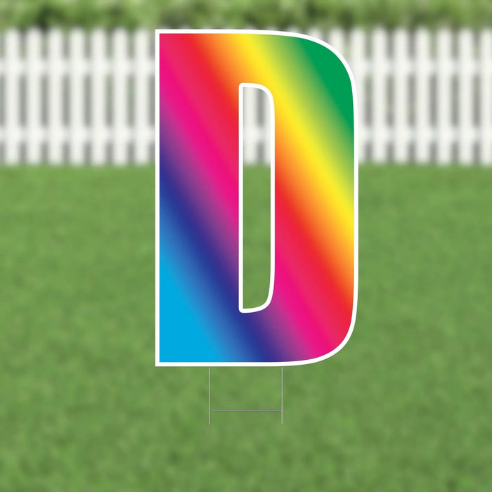 Colorful letter D yard sign with rainbow gradient from pink to blue, featuring a white outline and mounting stakes, displayed on grass with a white picket fence background.