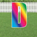 Colorful letter D yard sign with rainbow gradient from pink to blue, featuring a white outline and mounting stakes, displayed on grass with a white picket fence background.