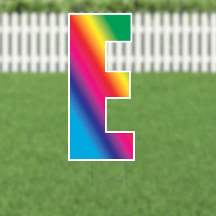 Colorful yard sign letter F with rainbow gradient fill and white outline, displayed on green grass with white picket fence background.